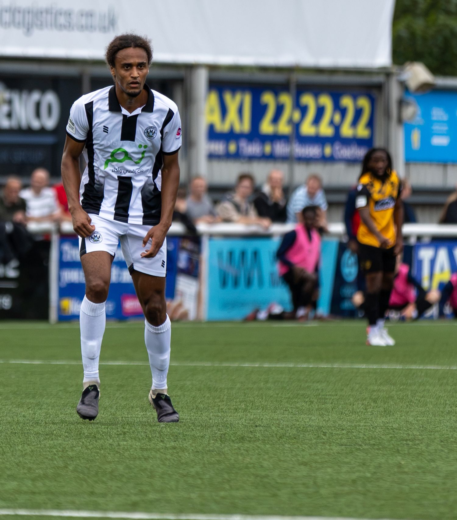 A footballer in black and white kit runs on a green pitch during a match, with advertising boards visible behind.