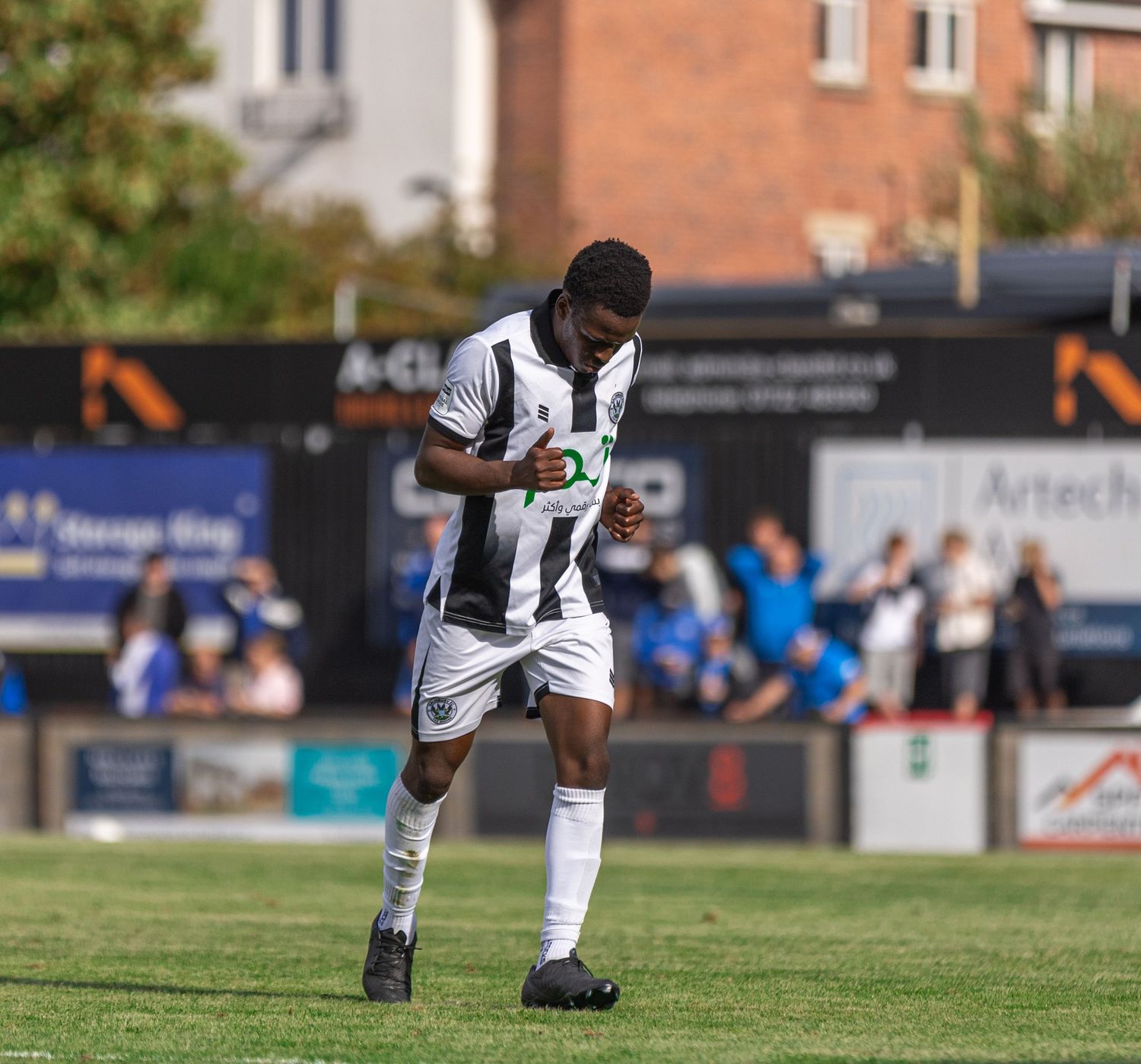 Soccer player in black and white striped kit celebrating on a sunny green pitch during a competitive match.