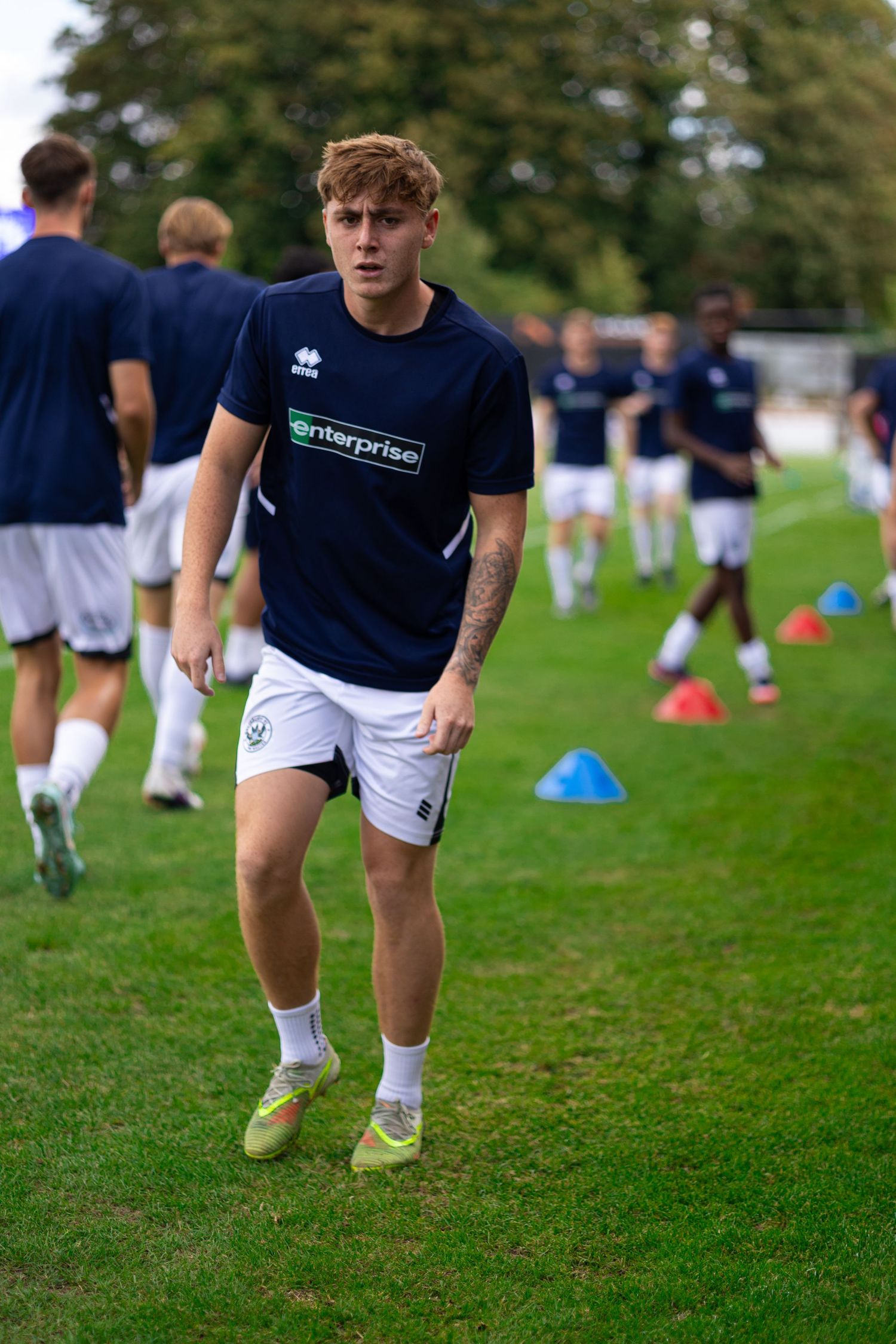 Young soccer player in navy Enterprise shirt and white shorts jogging during outdoor training drill with cones.