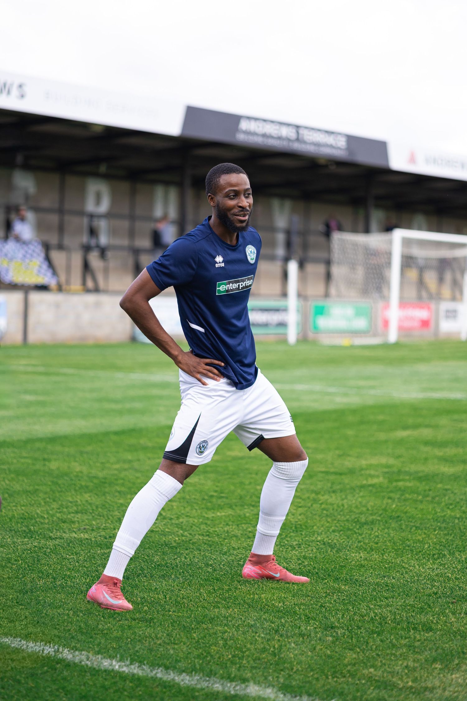 A smiling footballer in navy blue kit with white shorts and pink boots poses on a green pitch near a goalpost.