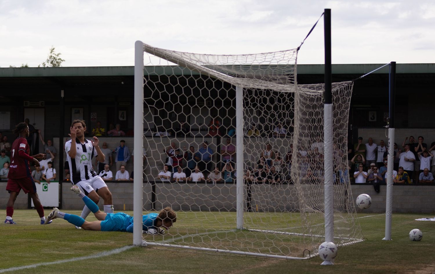 Goalkeeper lies on the ground after conceding a goal as players stand near the football net.