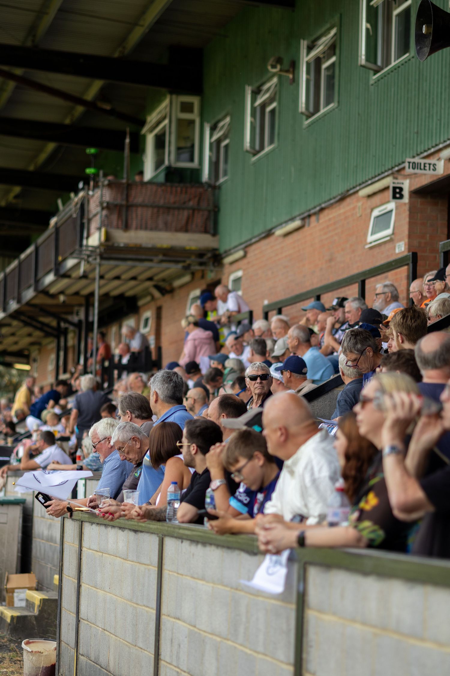 Crowds of football fans watching a match from the stands at a non-league ground with a green wooden grandstand.
