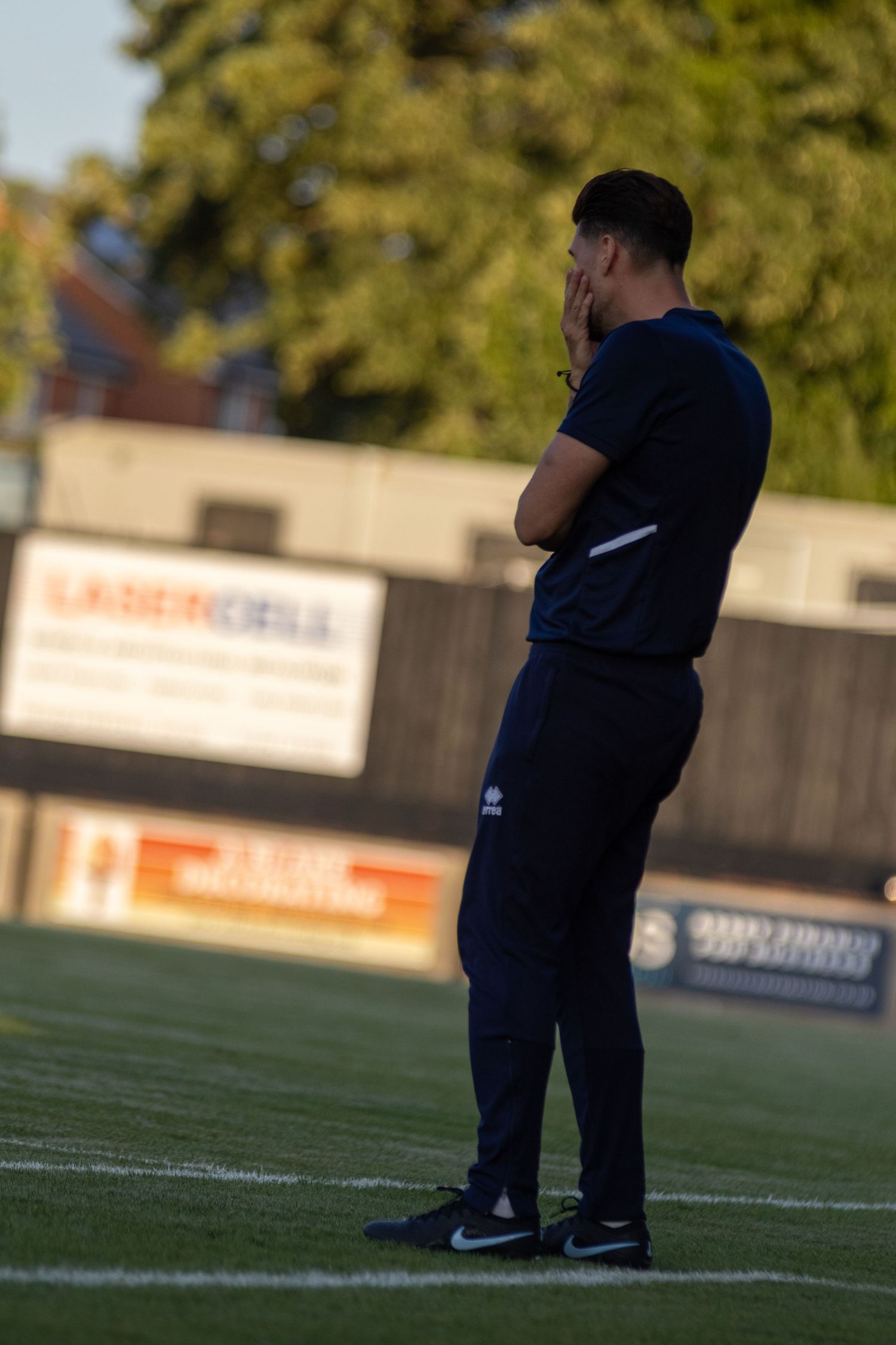 A football manager in dark navy tracksuit stands on the touchline watching a match intently.