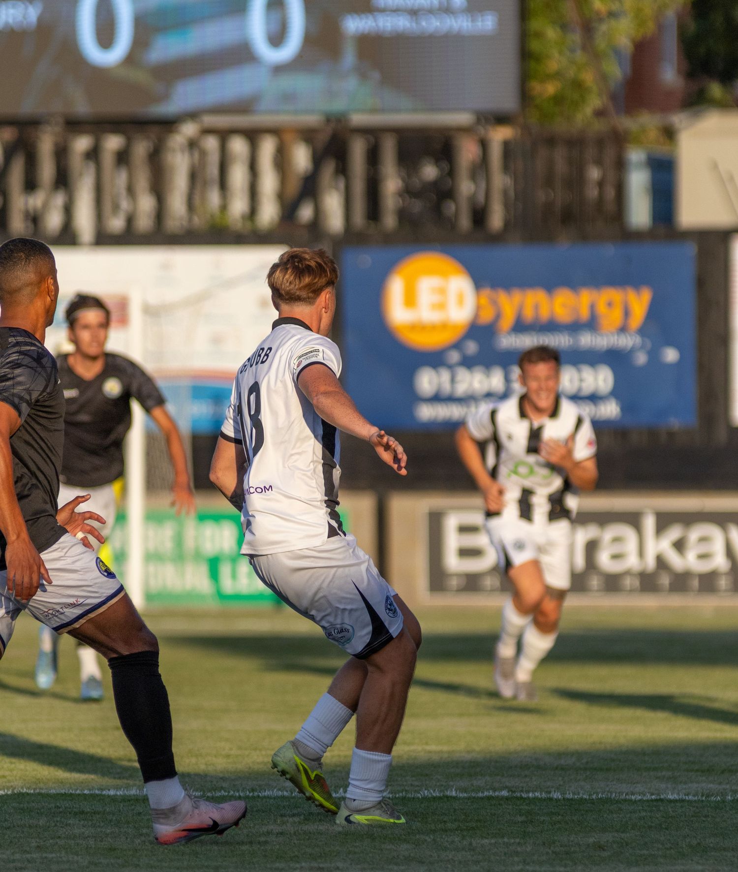 Soccer players compete during a match with a 0-0 scoreboard and LEDsynergy advertising board visible.