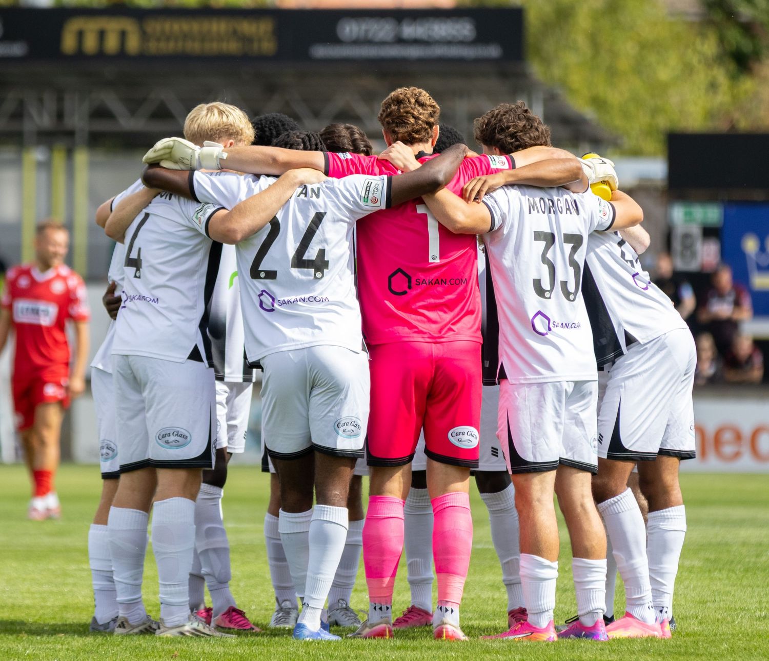 Football team huddles together on pitch before match in white and grey kit with goalkeeper in pink.
