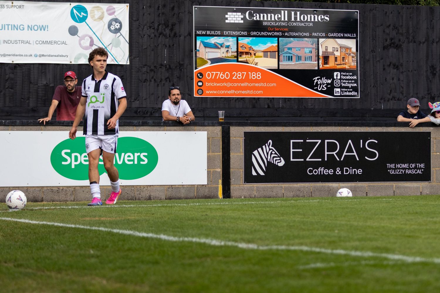 A footballer in black and white stripes prepares to take a corner kick at a non-league ground with advertising boards visible.