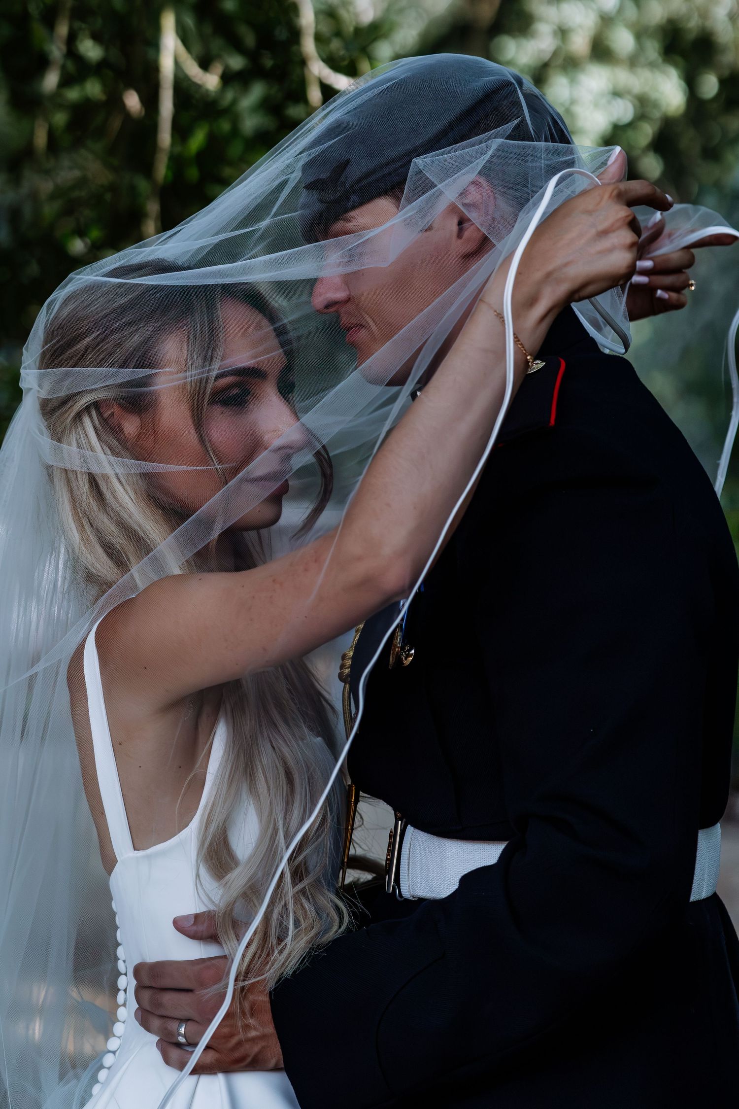 Bride and groom in military uniform sharing a romantic moment under a flowing wedding veil outdoors.