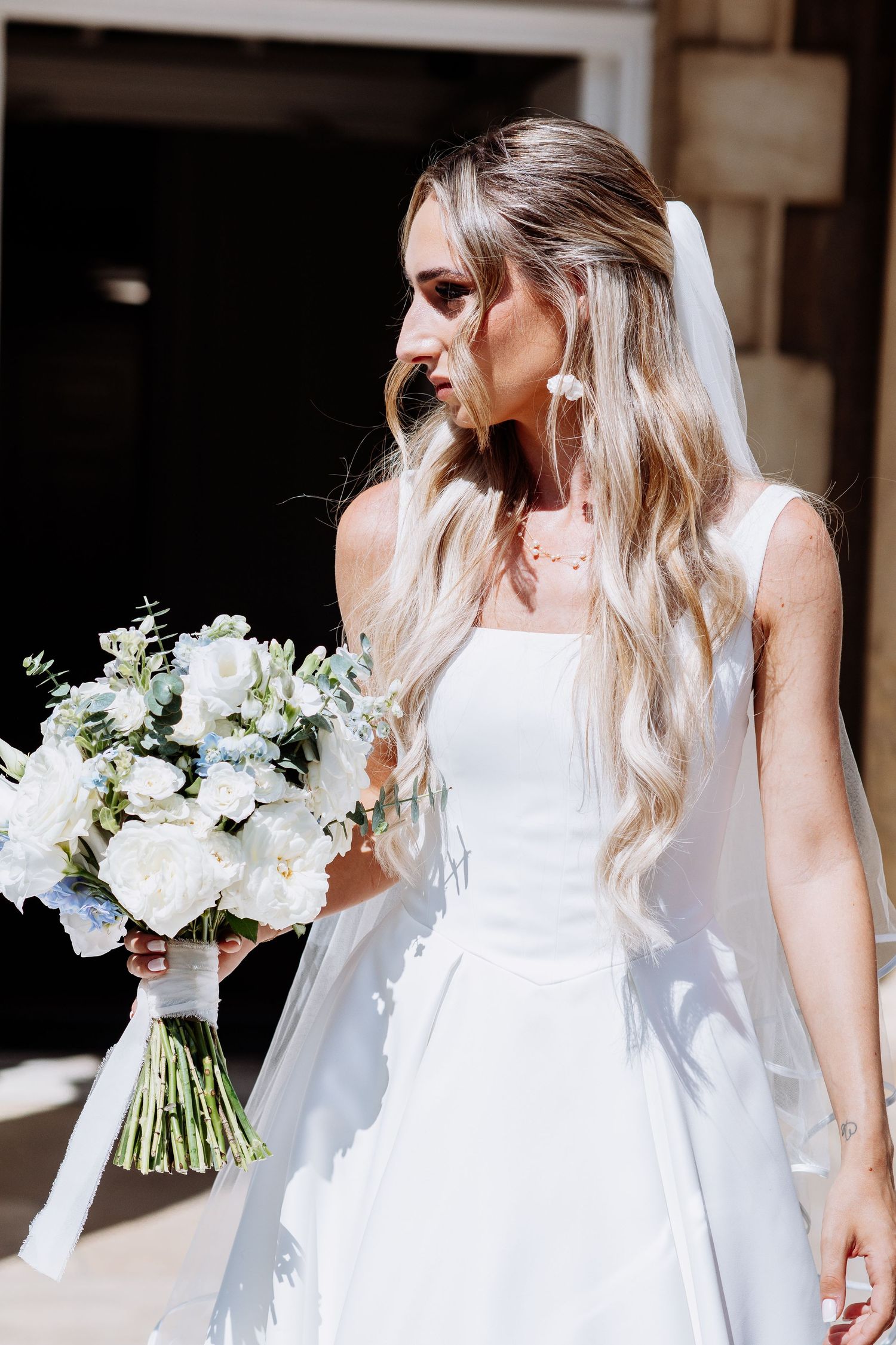 Stunning bride in white sleeveless gown with veil holds white and blue bouquet in bright sunlight.