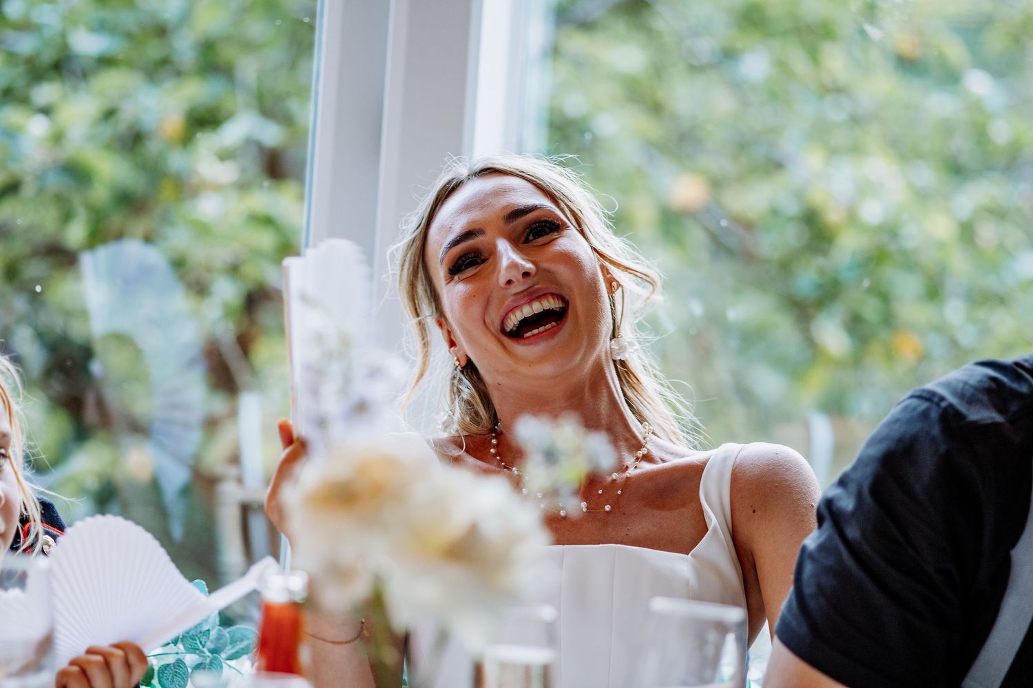 Bride laughing with bouquet at sunny wedding reception, wearing white dress and earrings.