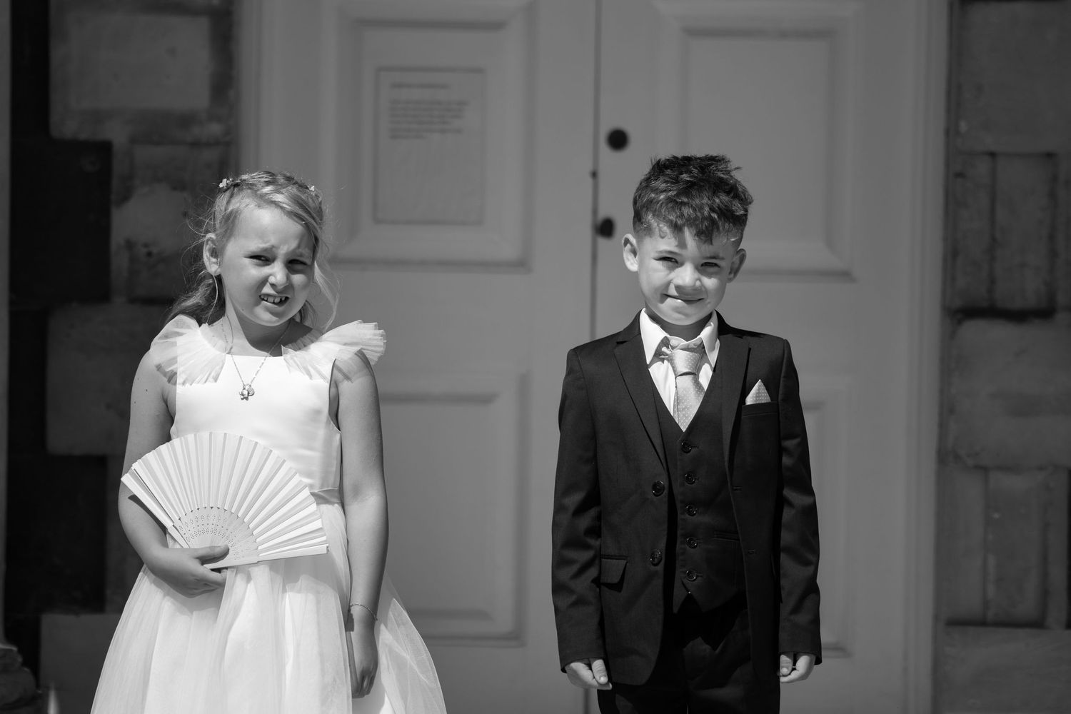 Black and white photo of a flower girl holding a fan and a young page boy standing together at a wedding.
