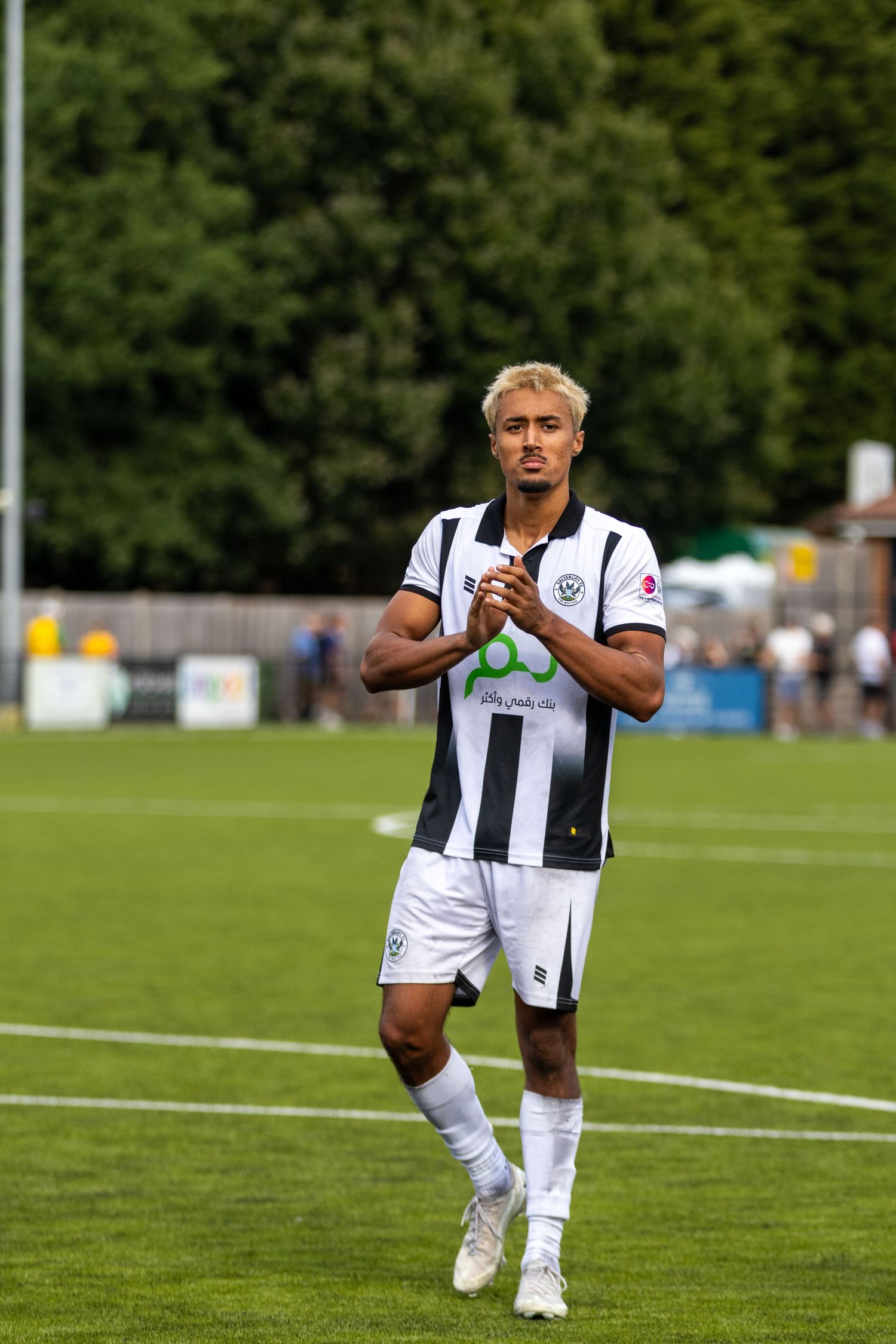 Blonde soccer player in black and white striped kit applauds while standing on a green football pitch.
