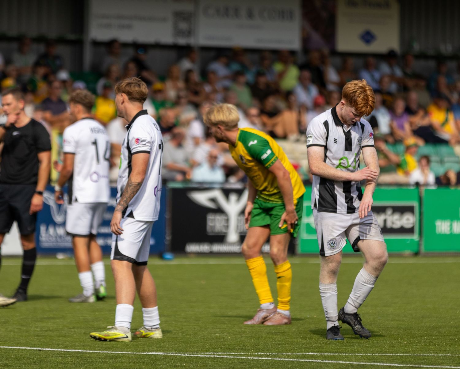 White-kitted players and a yellow-green opponent look dejected during a non-league football match on a sunny day.