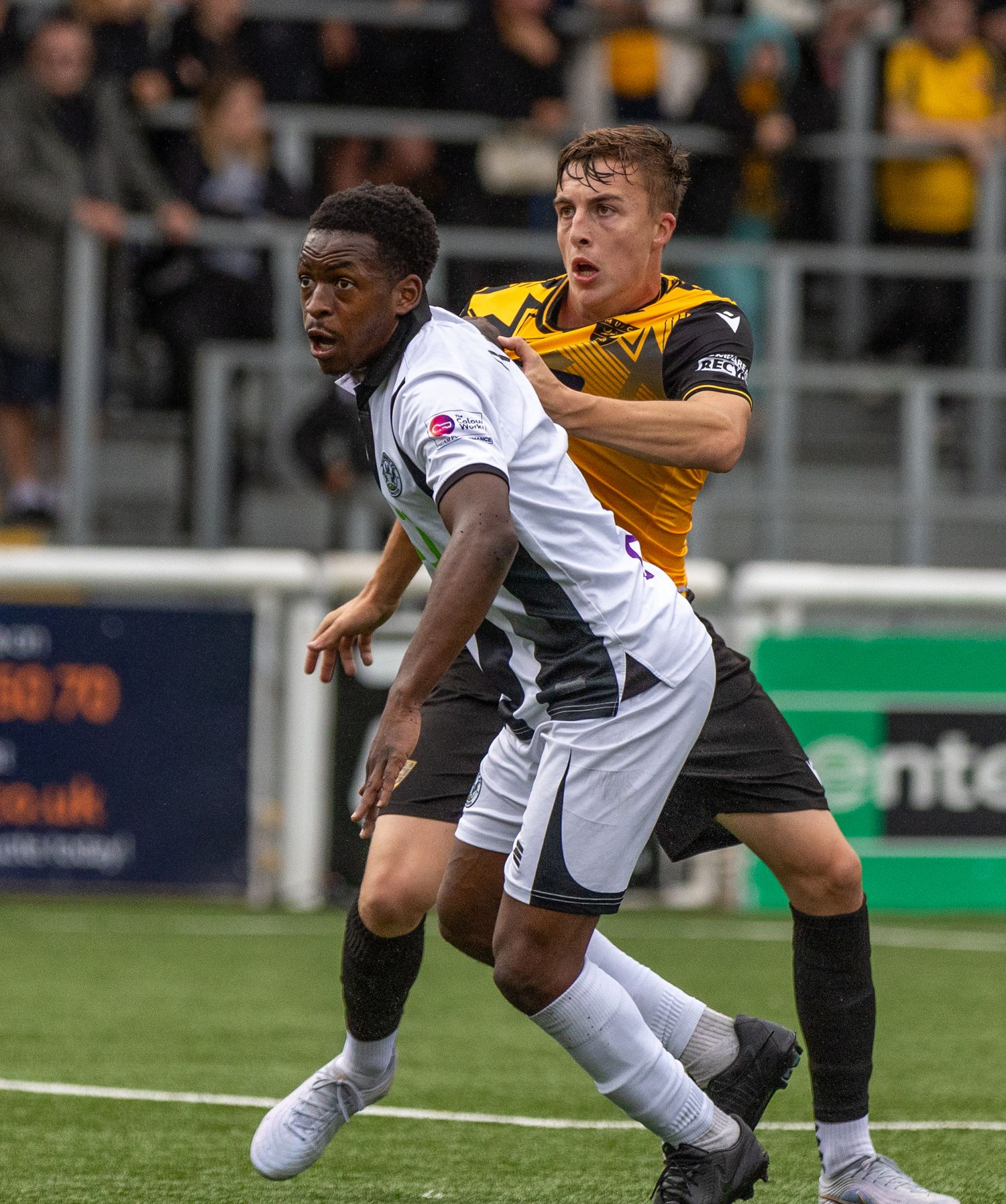 Two soccer players compete for the ball during a match, one in white and one in yellow and black kit.