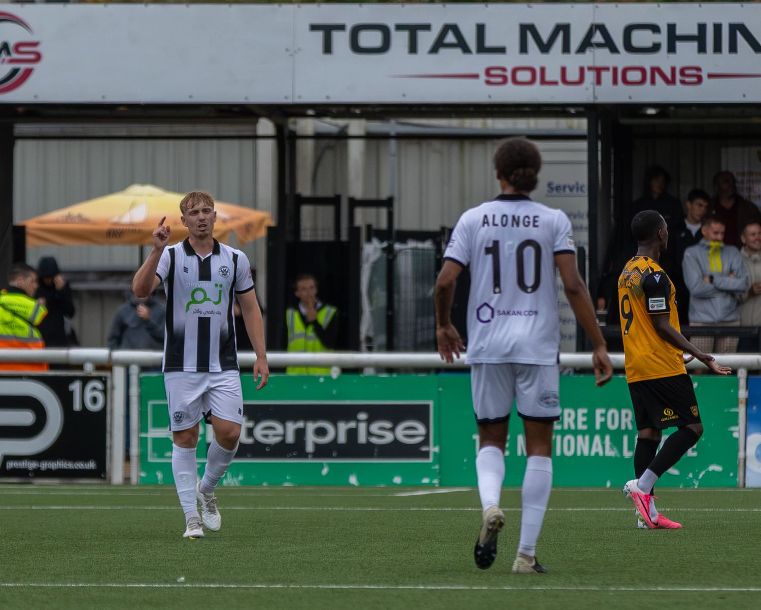 Football players in white jerseys, including number 10 Alonge, compete on a green pitch near Total Machine Solutions signage.