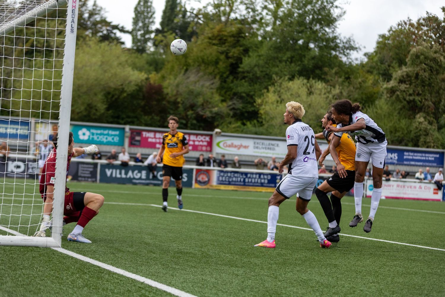 Soccer players compete in a header duel near the goal during an outdoor match on a green pitch.