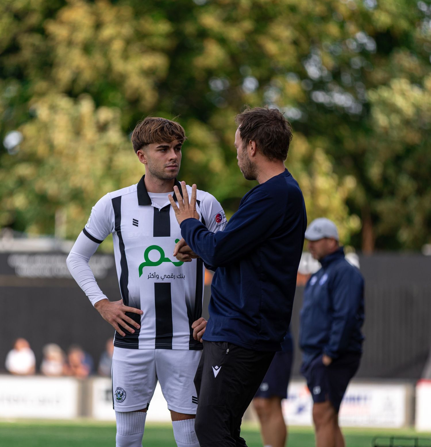 A football coach gives tactical instructions to a player in a black and white striped kit during a match.