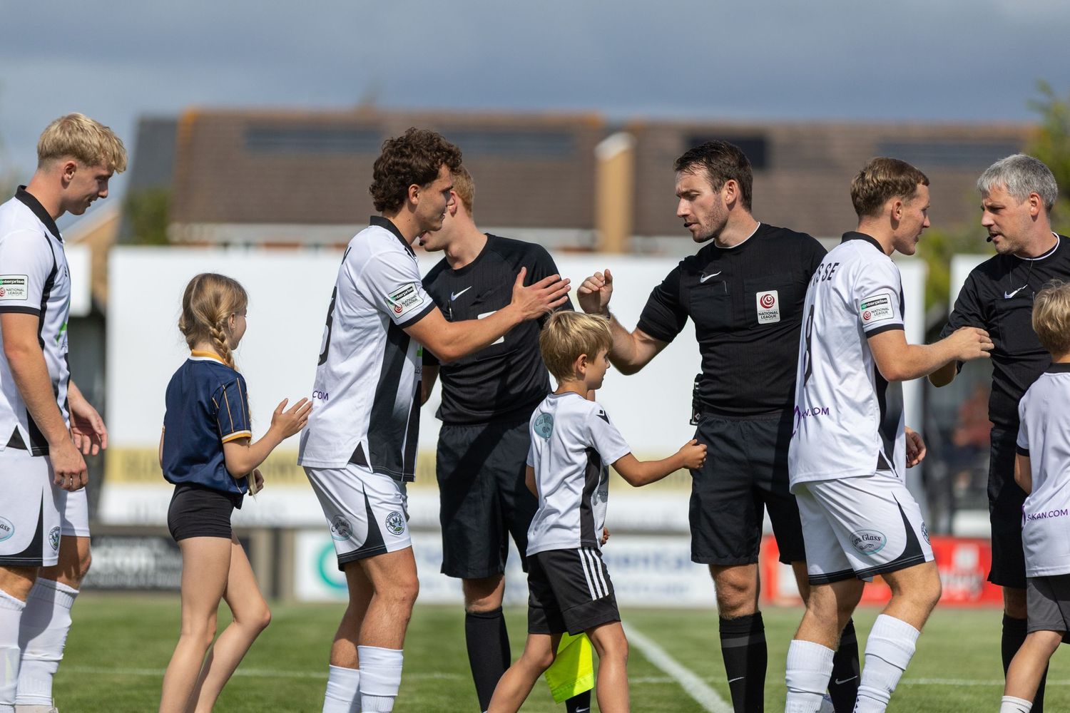 Football players and children shake hands before a match on a sunny day at a grass pitch.