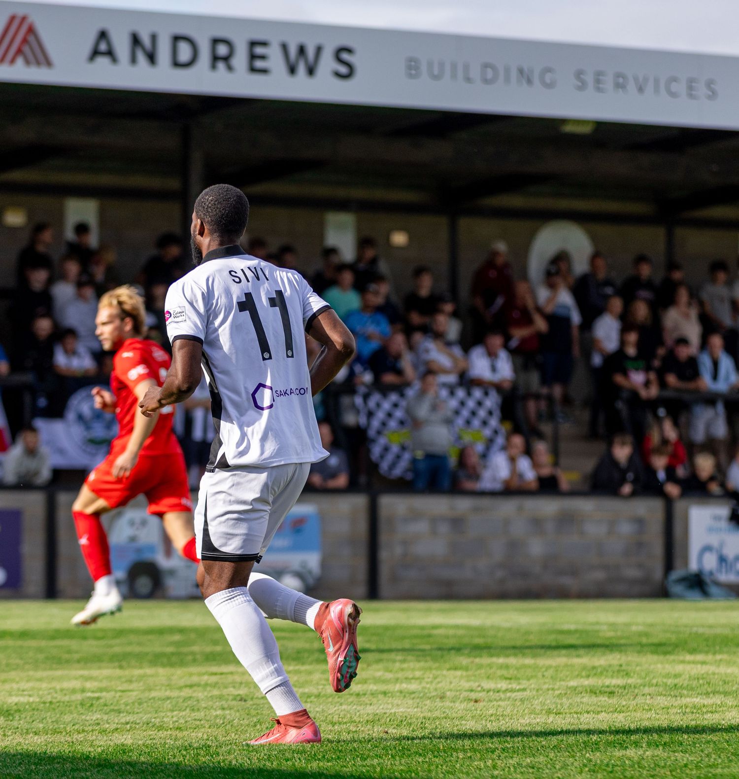 Player number 11 in white kit runs on green pitch at non-league football match, Andrews Building Services stand behind.