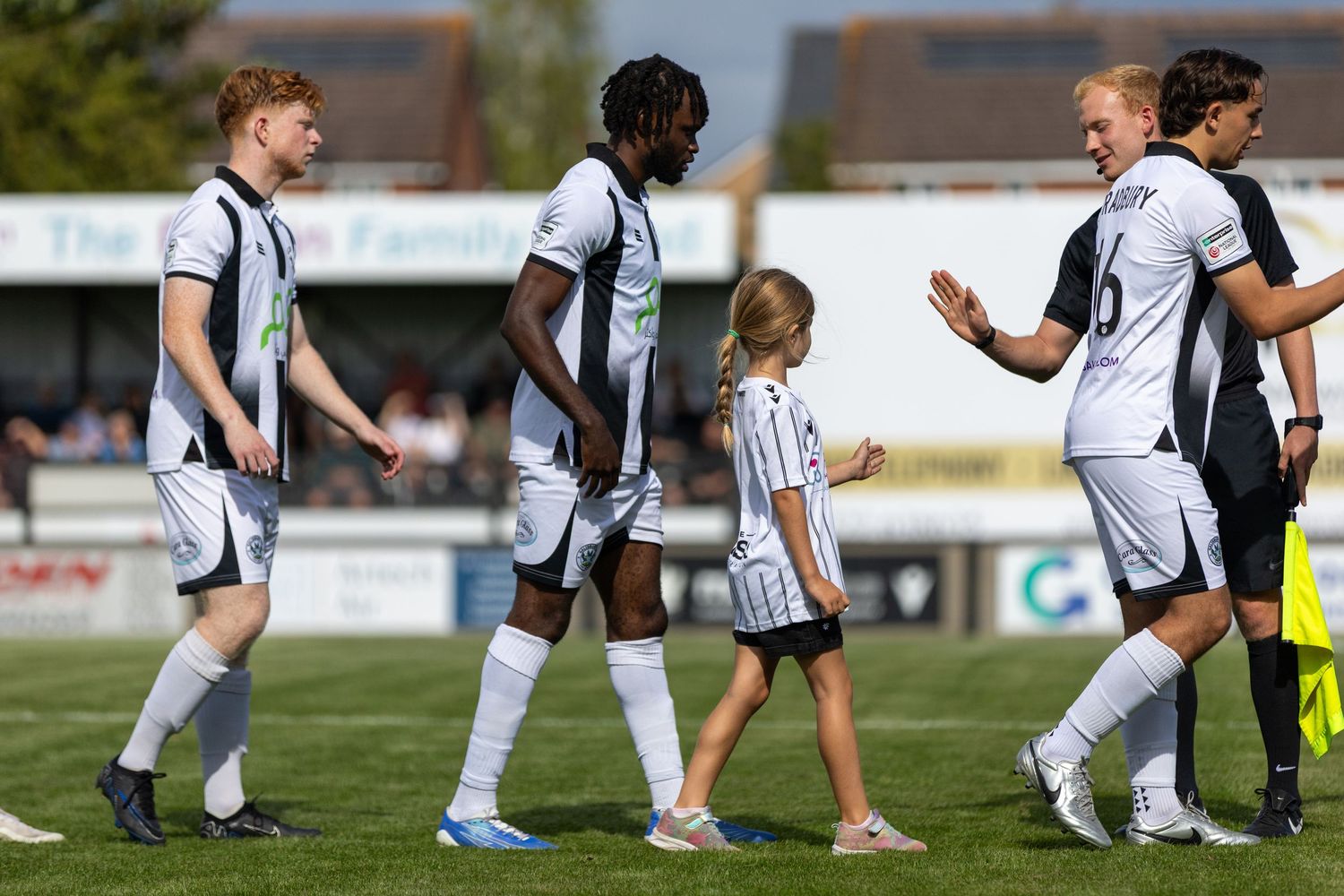 Football players in white uniforms walk onto a sunny pitch with a young child mascot before a match.