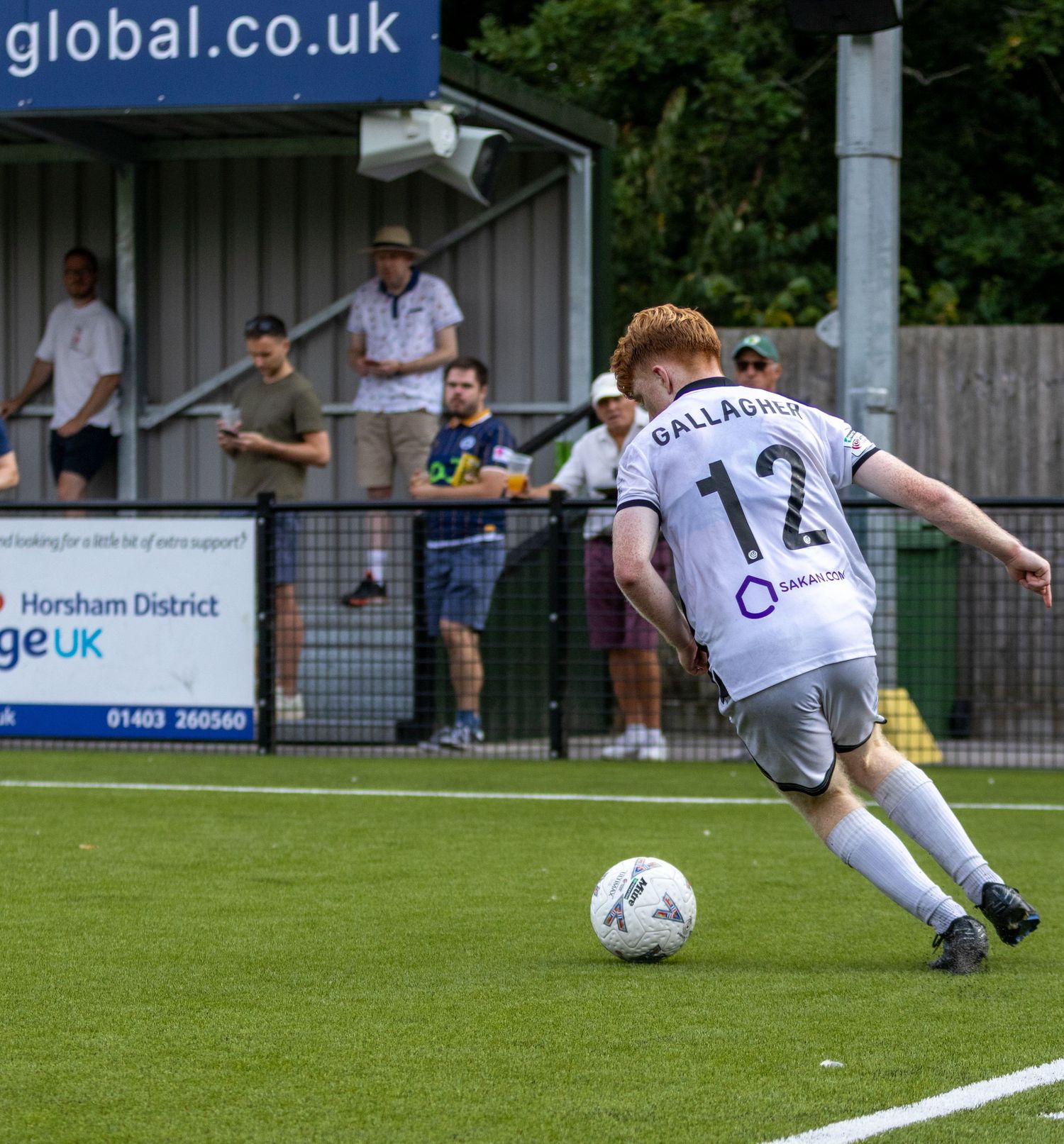 Gallagher number 12 in white kit strikes the ball near the touchline during a sunny football match.