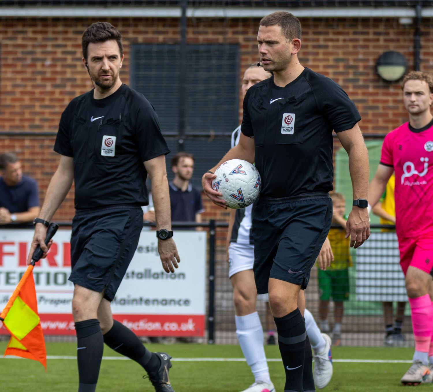Two football referees in black uniforms walk onto the pitch, one carrying a match ball during a non-league soccer game.