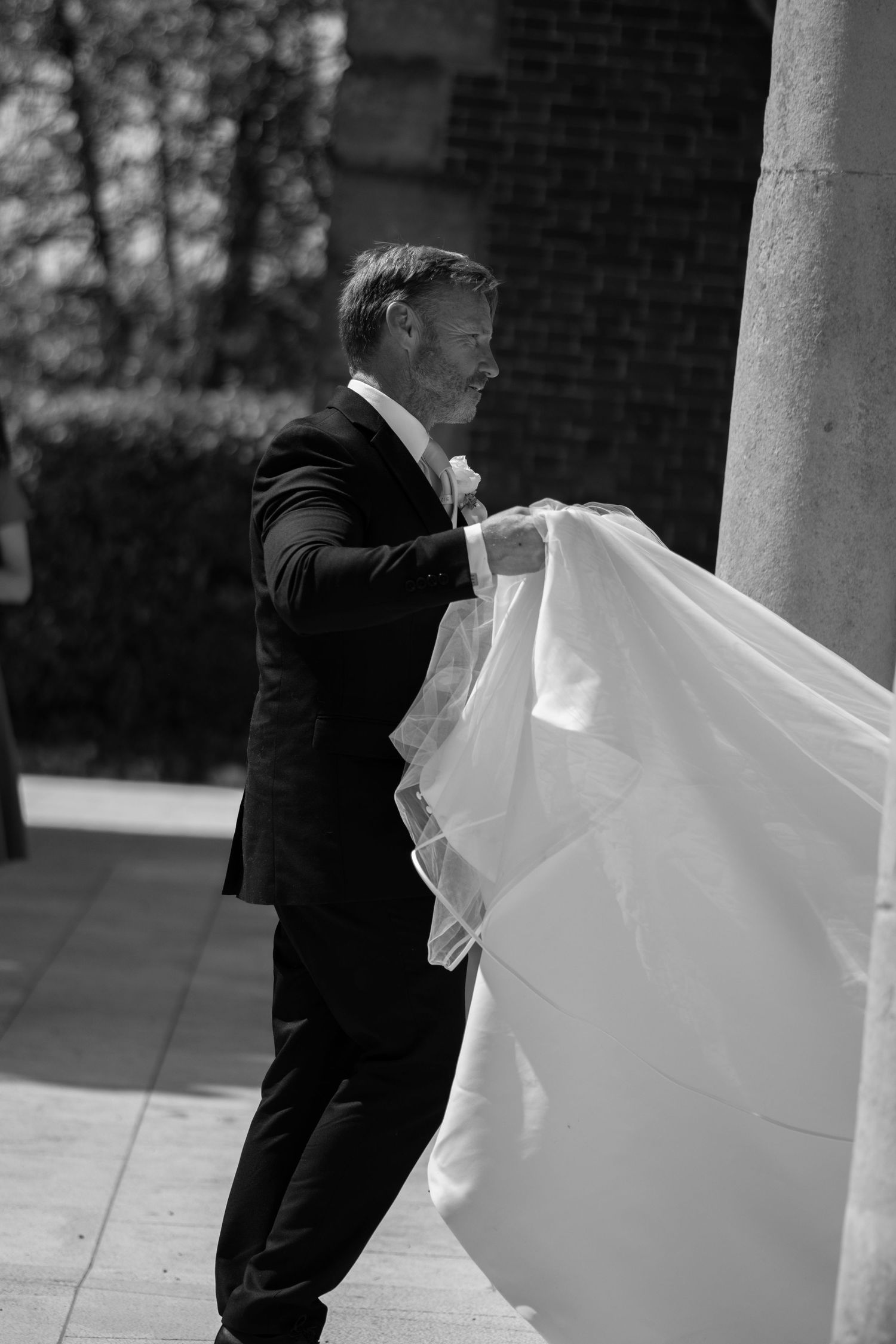 Black and white photo of groom in suit carefully arranging bride's wedding dress train outside.