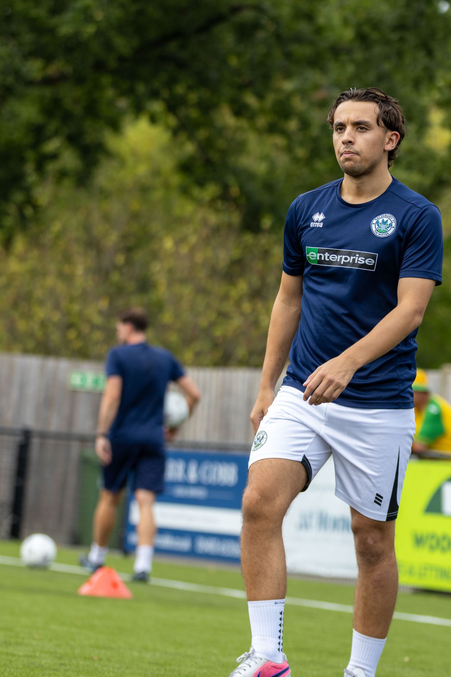 Male footballer in navy Enterprise-sponsored kit stretches legs during pre-match warmup on green pitch.