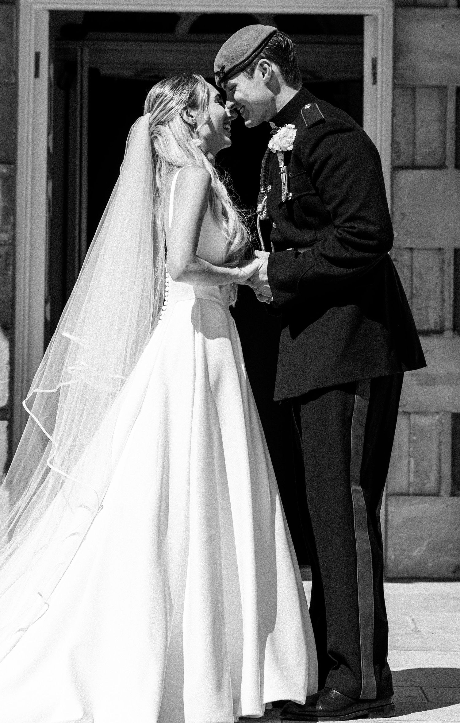 Black and white wedding photo of bride in flowing veil and groom in military uniform holding hands.