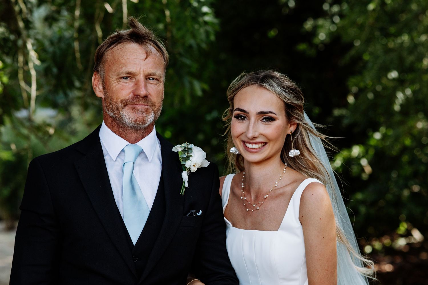 Older man in black tuxedo with blue tie stands beside smiling bride in white dress at outdoor wedding.