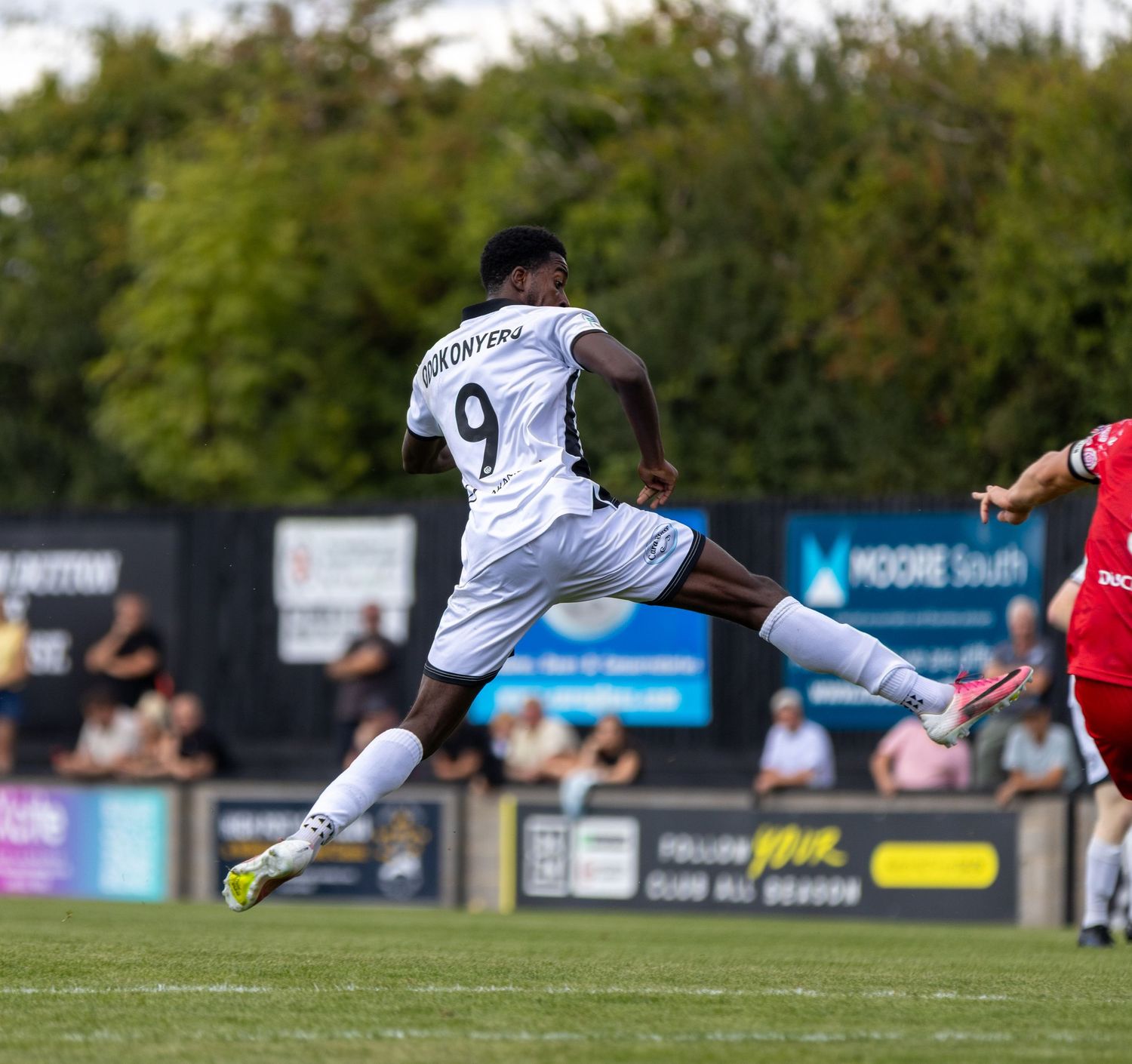 Odokonyero number 9 in white kit leaps dynamically to kick the ball against a red-shirted opponent on green pitch.