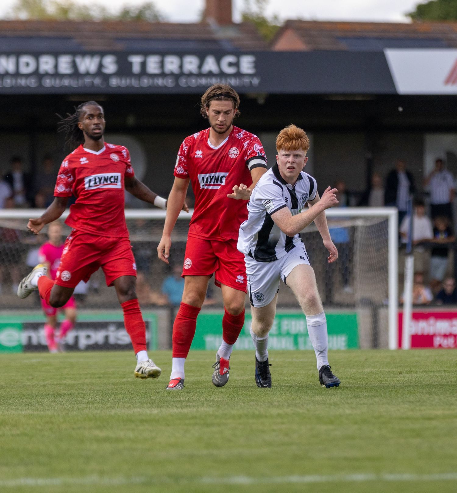 Two red-kitted players chase a young ginger-haired player in black and white kit at Andrews Terrace ground.