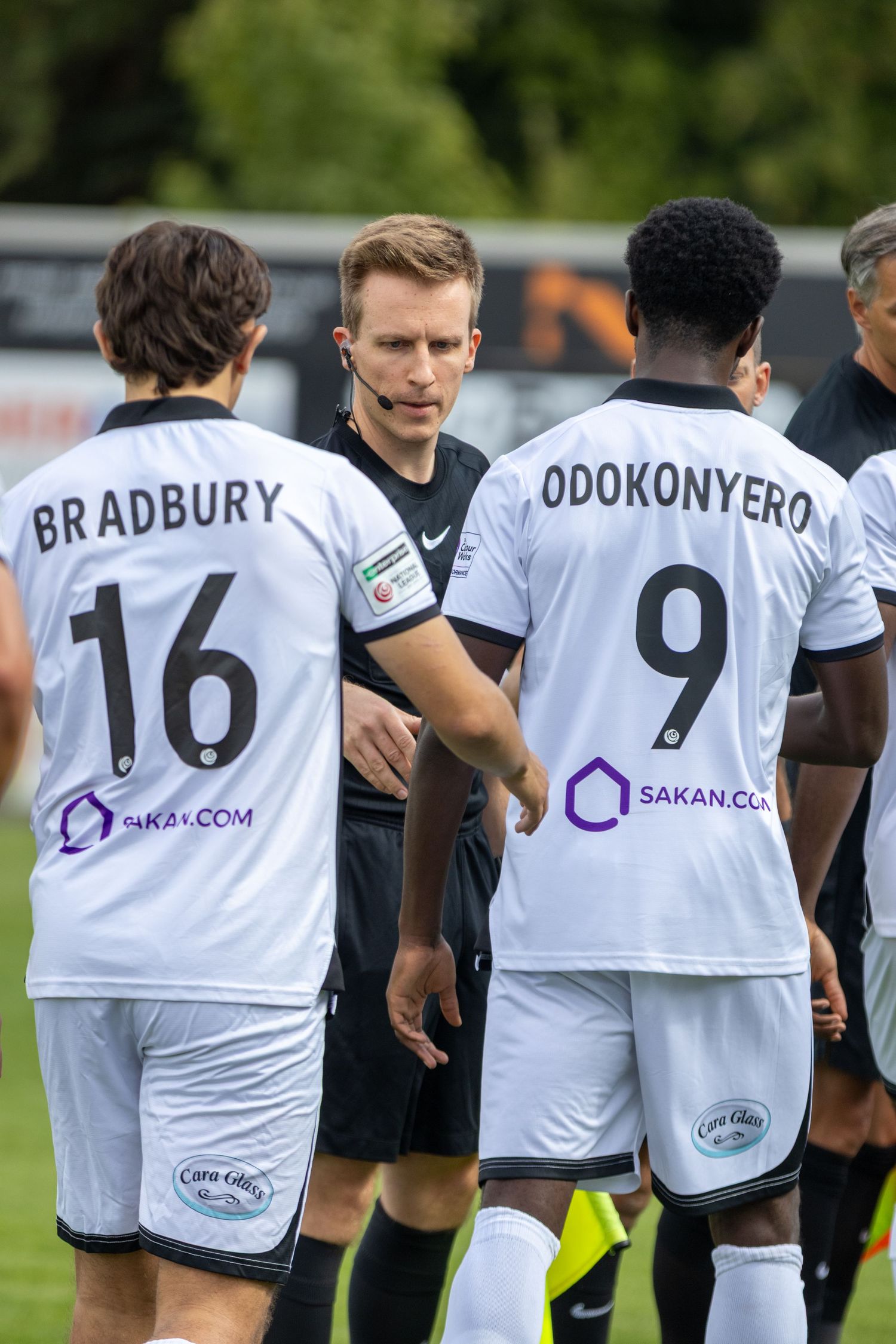 Referee speaking to Bradbury 16 and Odokonyero 9 in white football kits during a match.