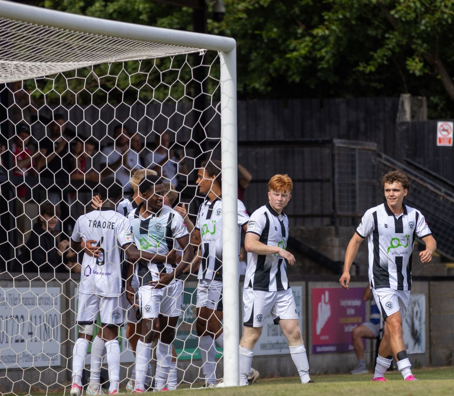 Salisbury FC players gather to celebrate a goal near the goalpost during a sunny home football match.