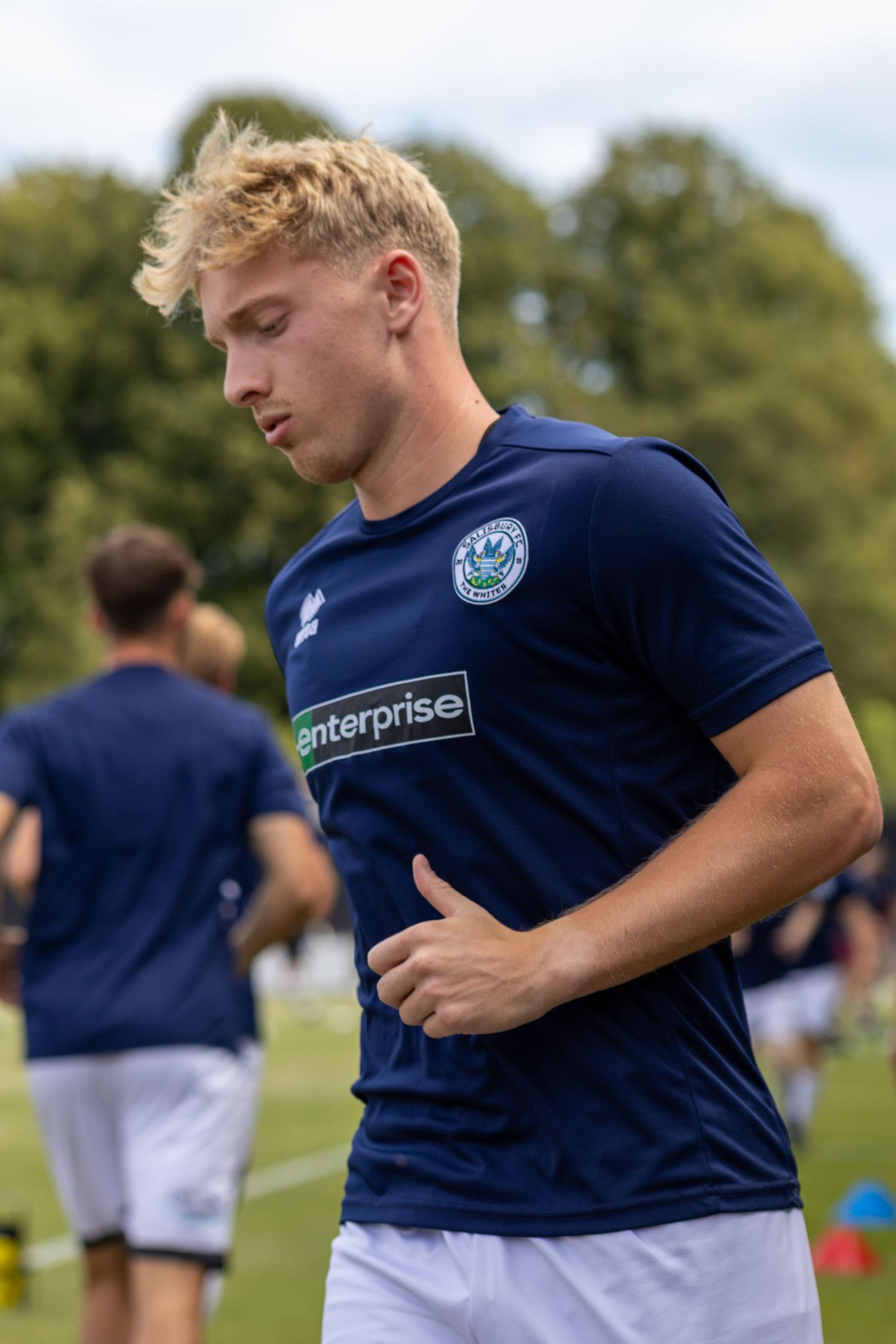 Young blonde footballer in navy Enterprise-sponsored shirt jogging during pre-match warmup on grass pitch.
