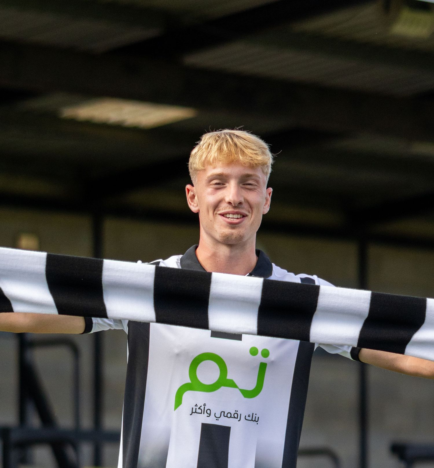 A smiling blonde soccer player proudly holds up a black and white striped jersey with arms outstretched.