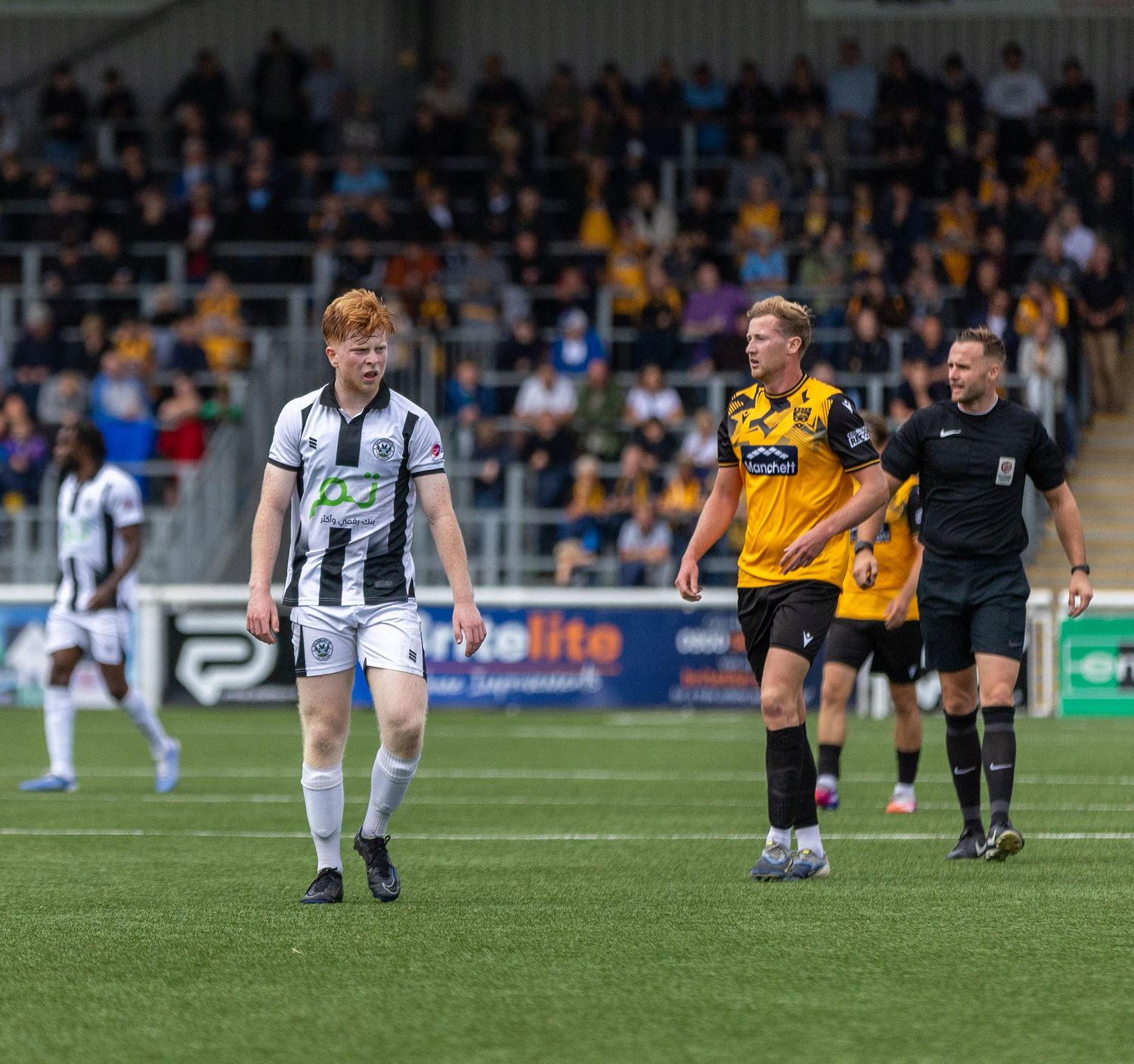 Football players and referee walk on pitch during a match, with fans in the stadium stands behind them.