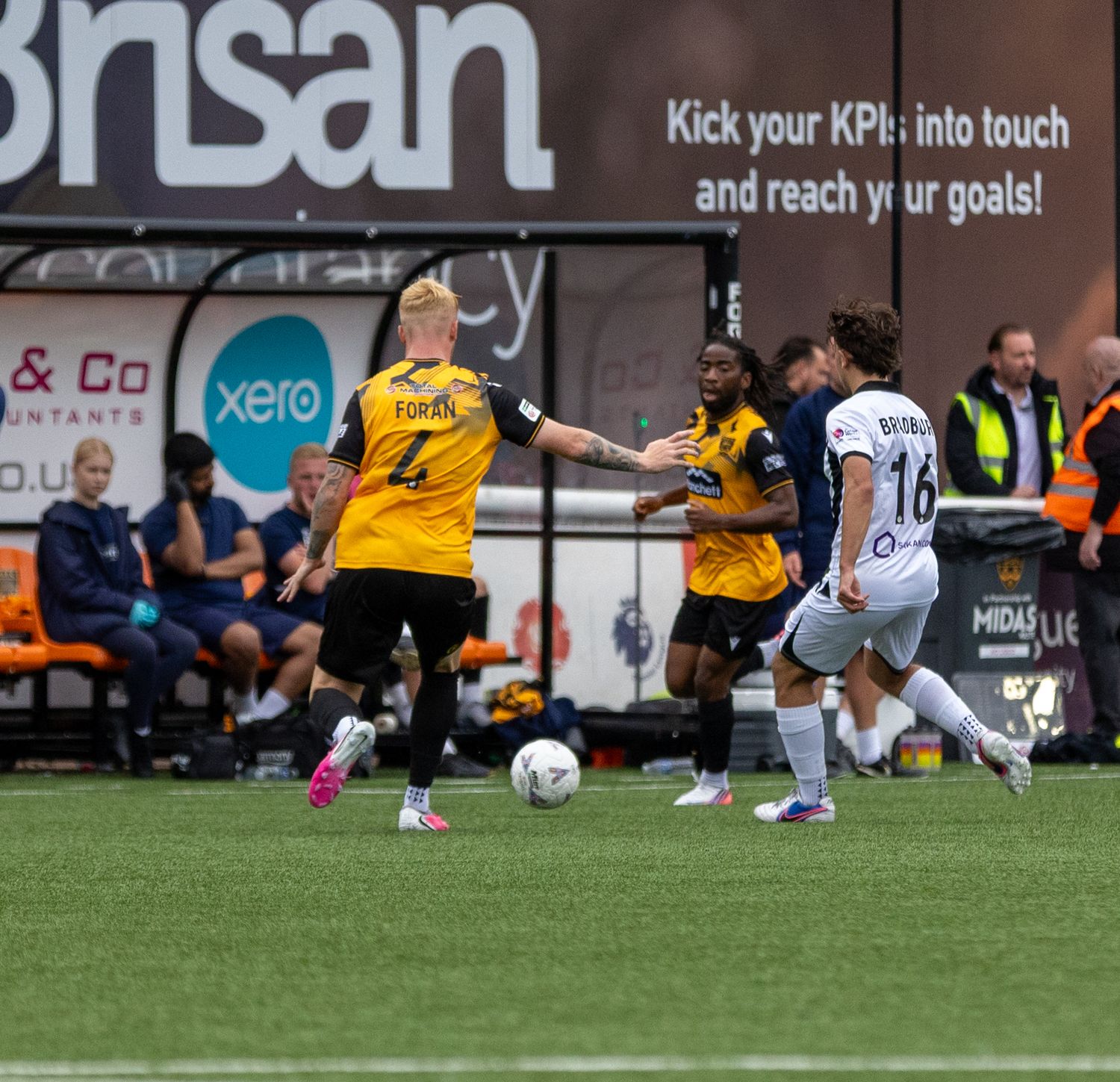 Soccer players compete for the ball during a match, with yellow and white kits visible on a green pitch.