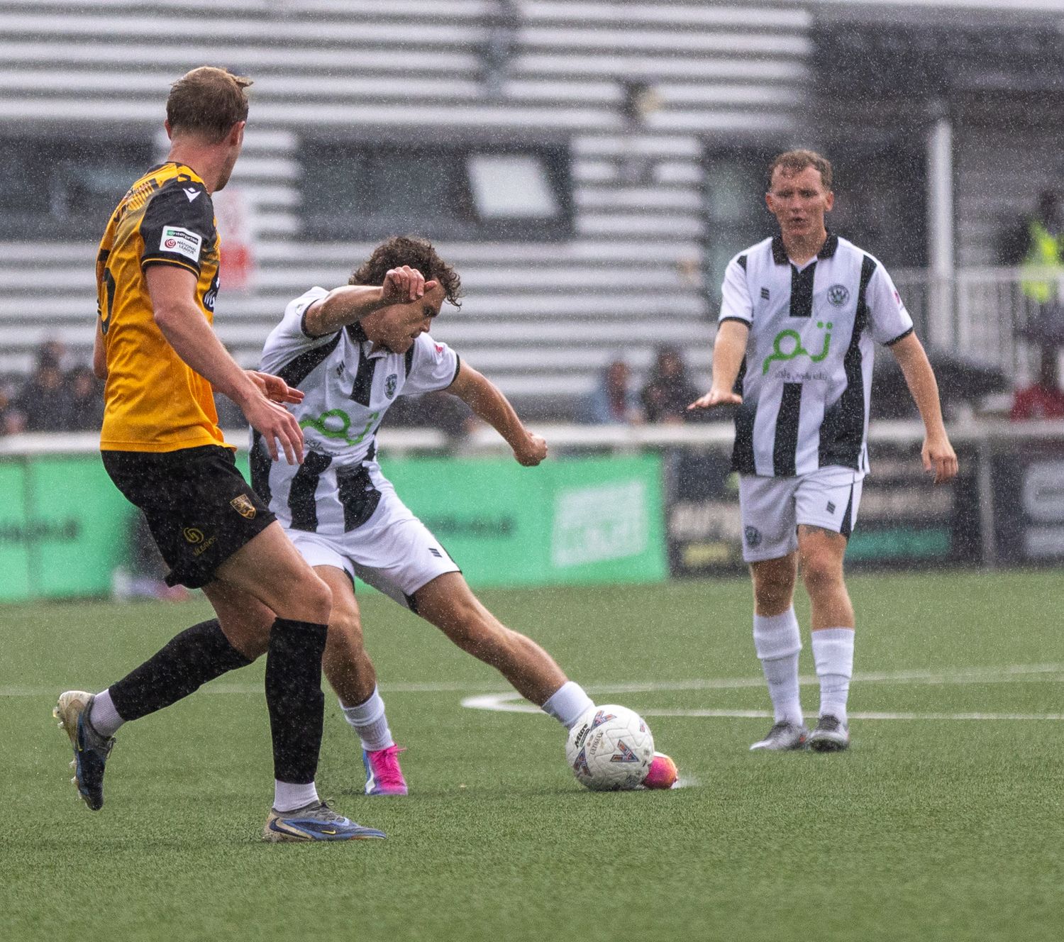 Soccer players in black-and-white and yellow kits compete for the ball on a green pitch during a match.