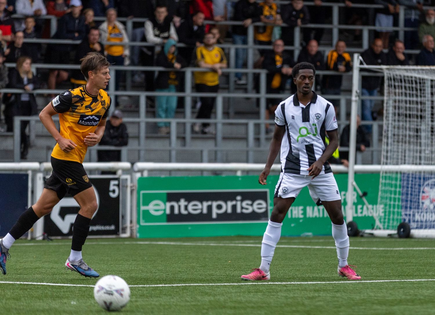Two soccer players compete on a green pitch during a National League match, with an Enterprise sponsor banner visible.
