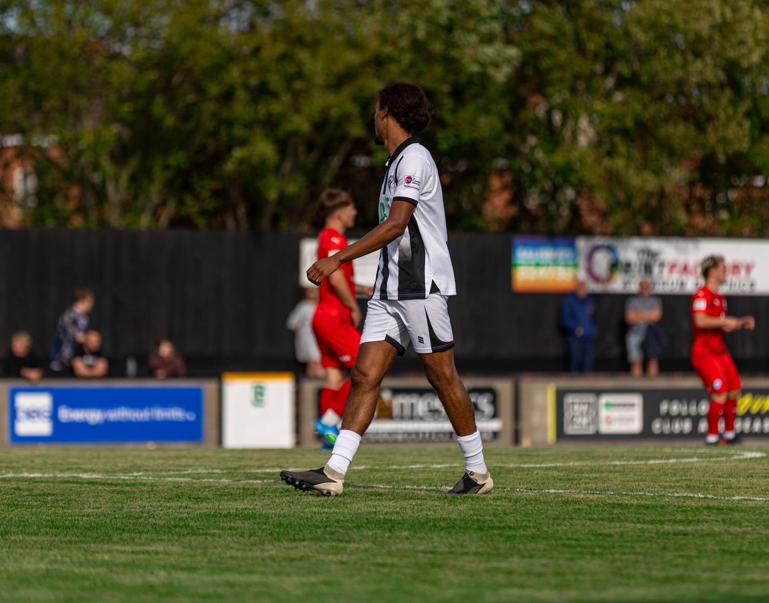A soccer player in a black and white kit walks on a green pitch during a sunny match with red-uniformed opponents in background.