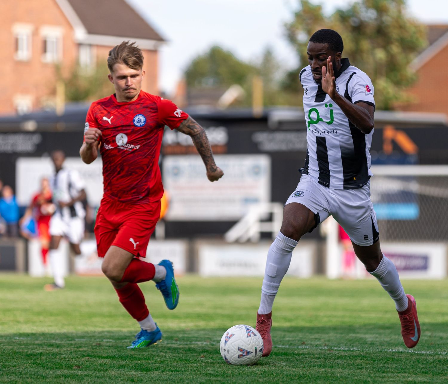 Two soccer players compete for the ball during a match, one in red and one in black and white striped kit.