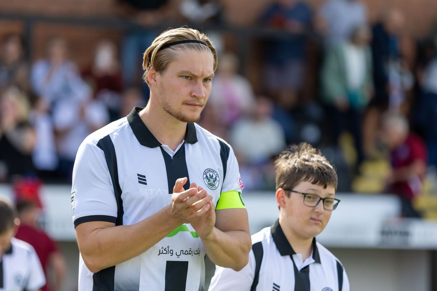 Football player in black and white kit clapping on pitch with young mascot beside him during a match.