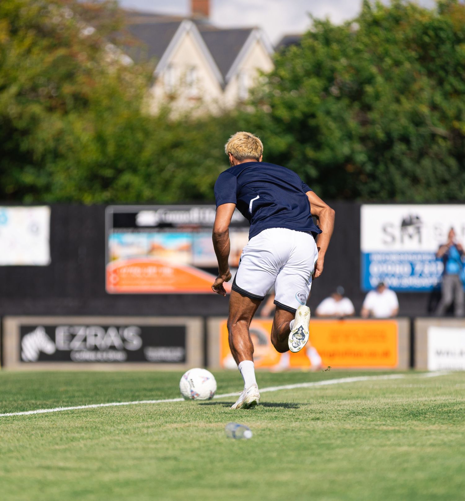 A blonde soccer player in navy blue shirt and white shorts dribbling a ball on a green field during a match.