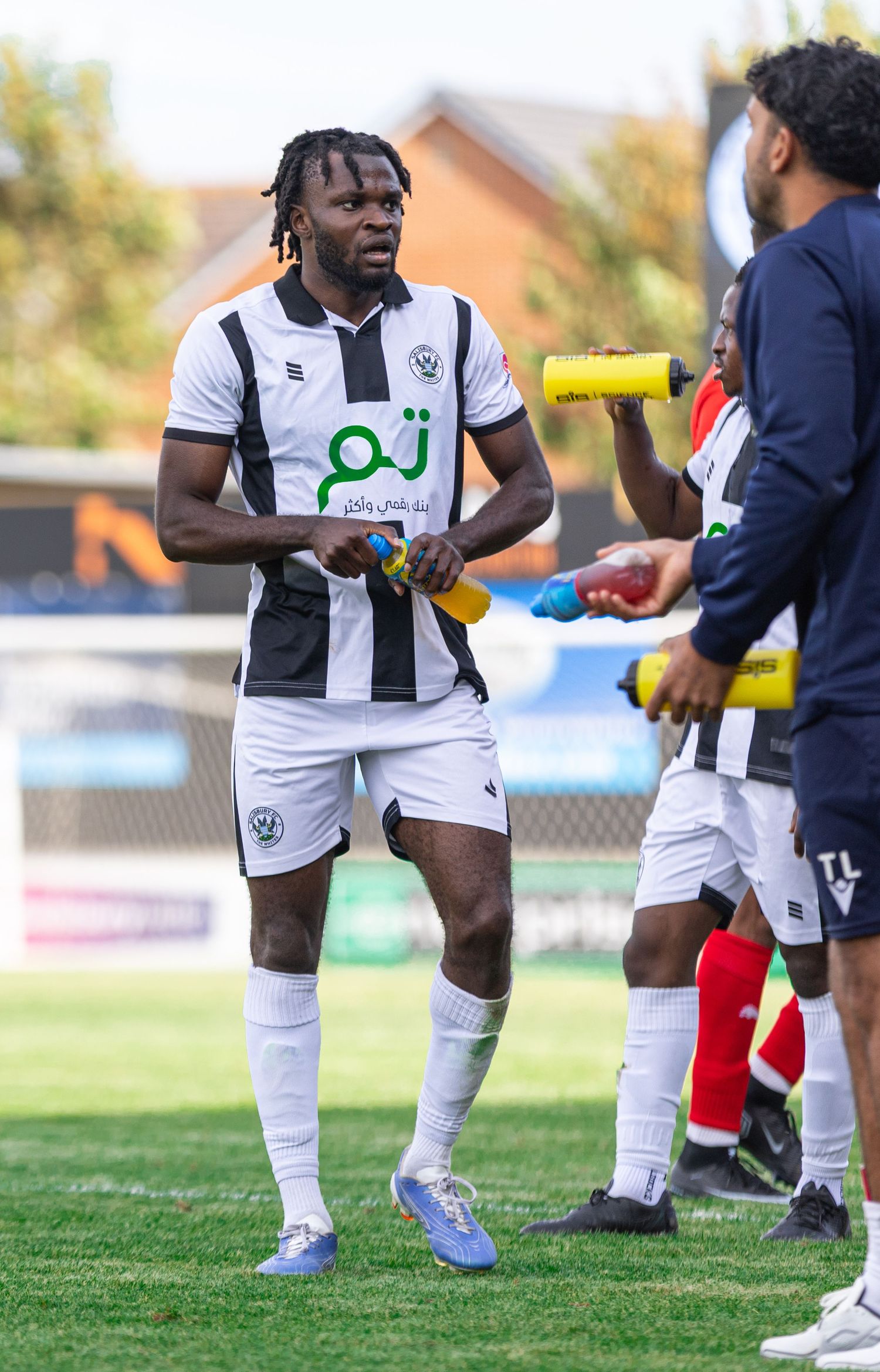 A Black male soccer player in white and black kit holds a water bottle on the field during a match break.
