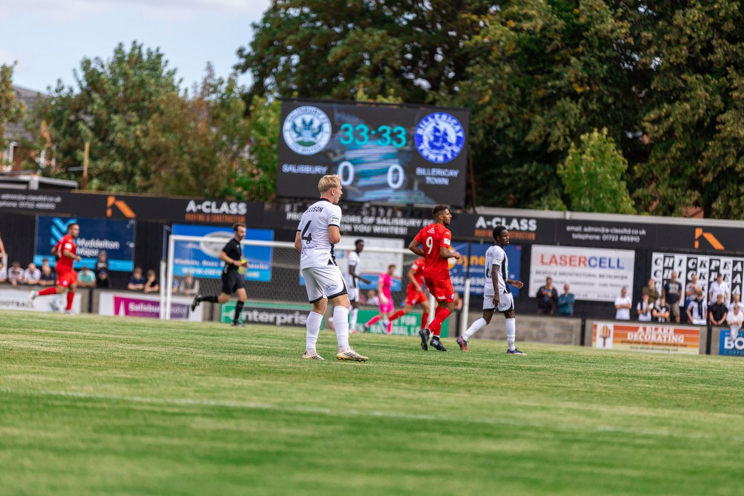 Soccer players compete on a green field during a match, with a scoreboard showing 0-0 at 33:33 in sunny weather.