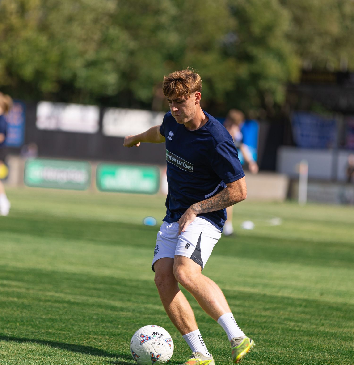 Young male soccer player in navy kit dribbling a ball on green grass pitch during a sunny outdoor training session.