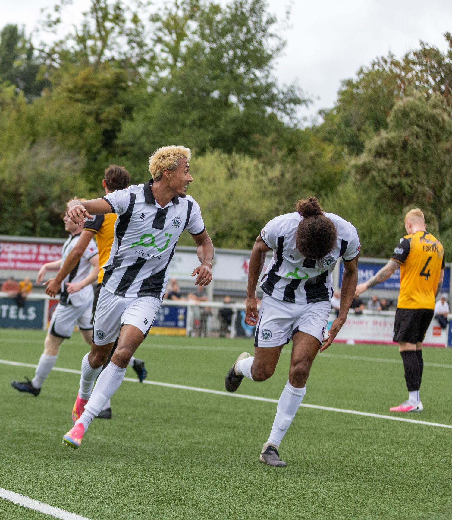 Two soccer players in white uniforms celebrate a goal during a match on a green field with fans in the background.