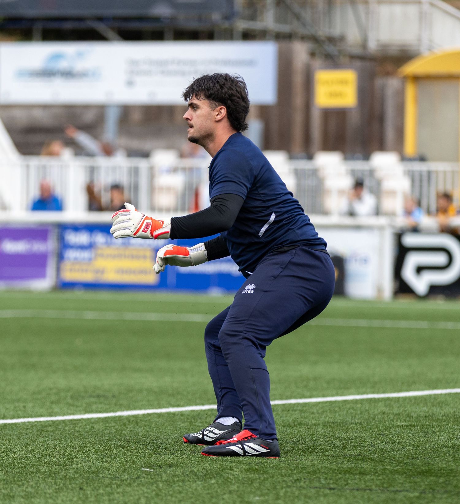 Goalkeeper in navy kit crouches in ready position on football pitch during match.