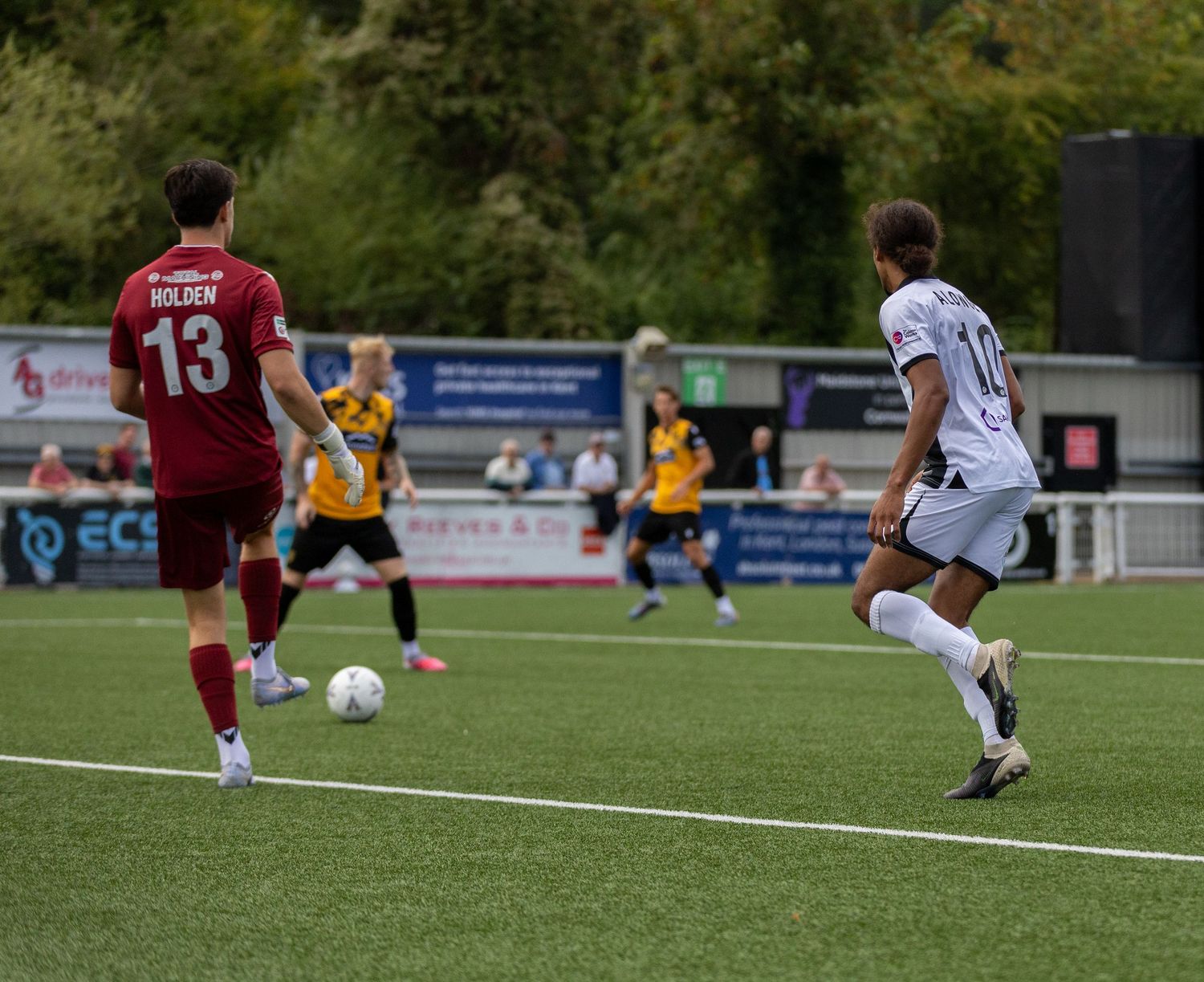 Soccer players compete on a green turf field, with player number 13 Holden in red visible during a sunny match.