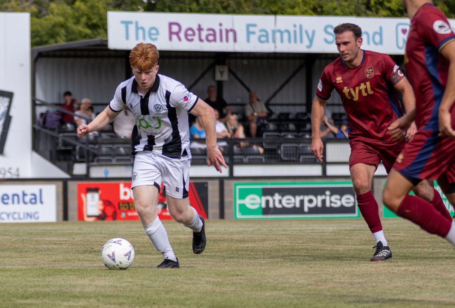 A red-haired footballer in black and white strip dribbles the ball during a sunny non-league football match.